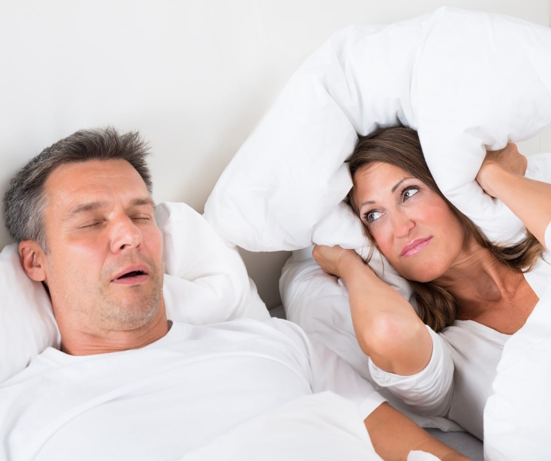 annoyed woman holding pillow over her ears as husband lays next to her snoring